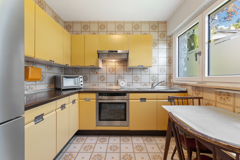 Gelbe Einbauküche mit Backofen, Toaster, Tisch und zwei Stühlen, Fliesen mit Blumenmuster.