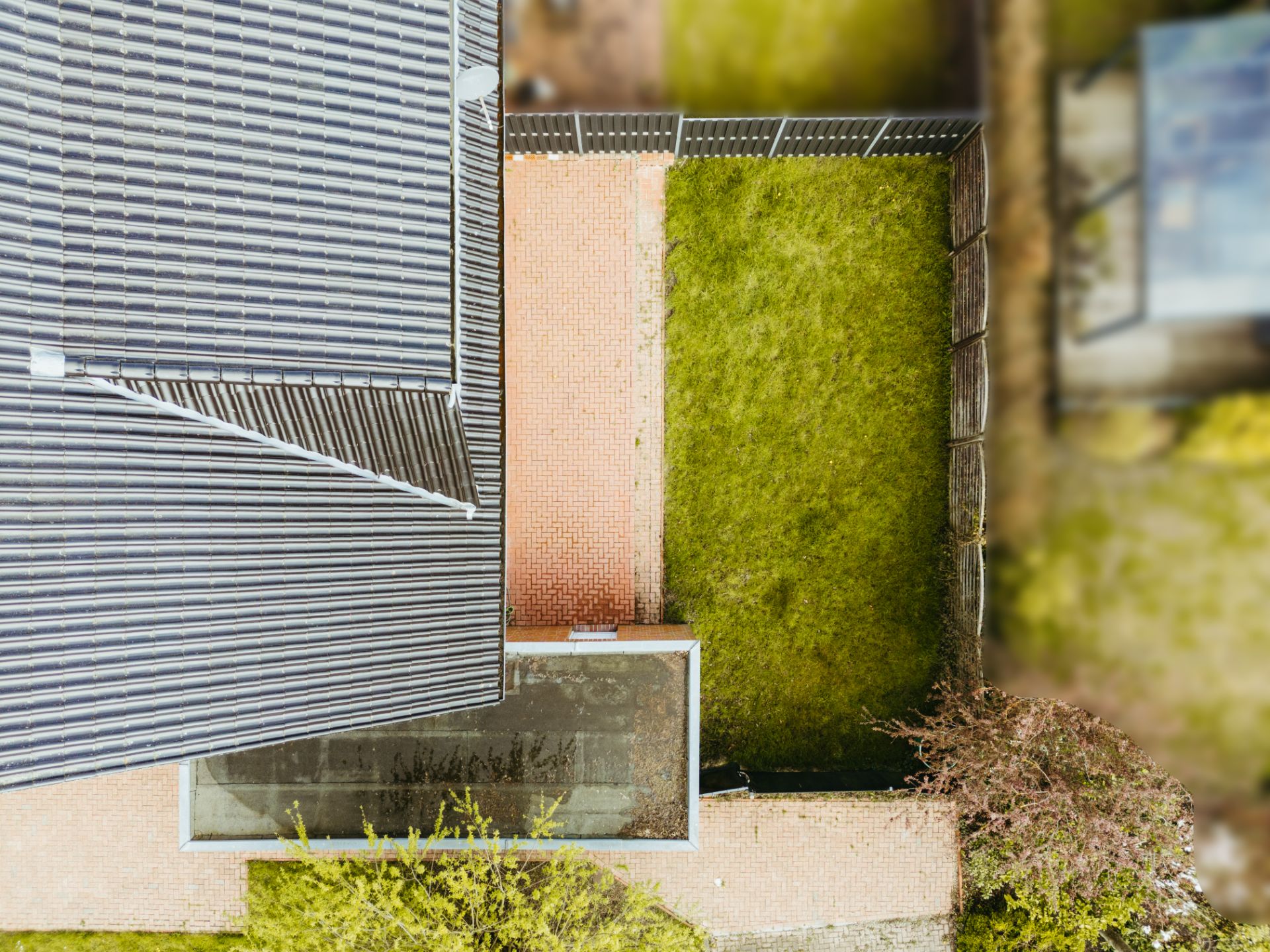 Luftaufnahme eines Hauses mit angrenzendem Garten und gepflasterter Terrasse, umgeben von einem Zaun.