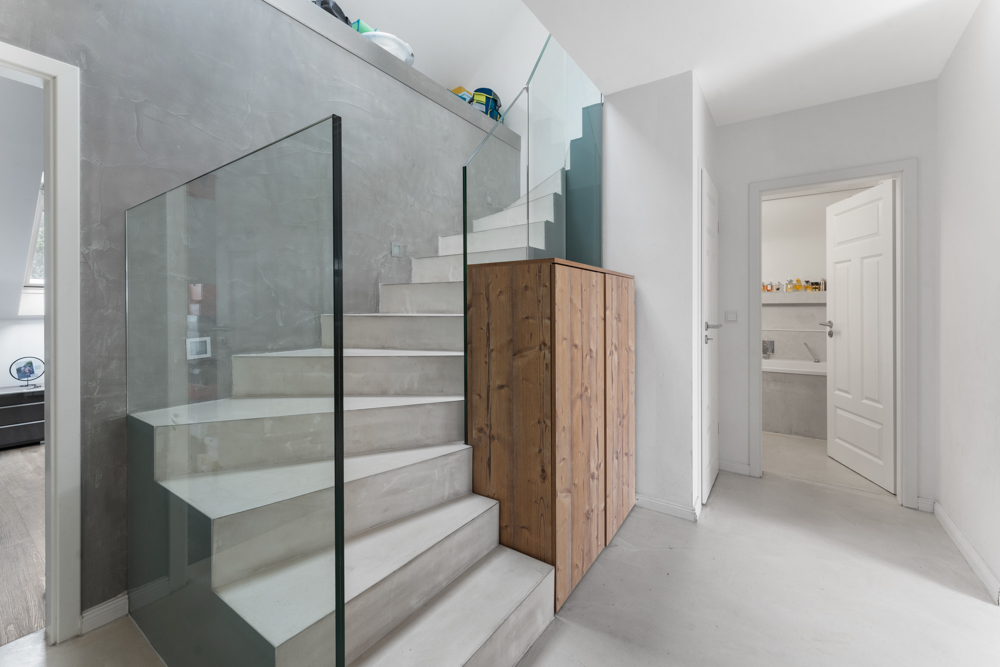 Moderne Treppe mit Glasgeländer und Holzschrank in einem hellen Flur mit zwei offenen Türen.