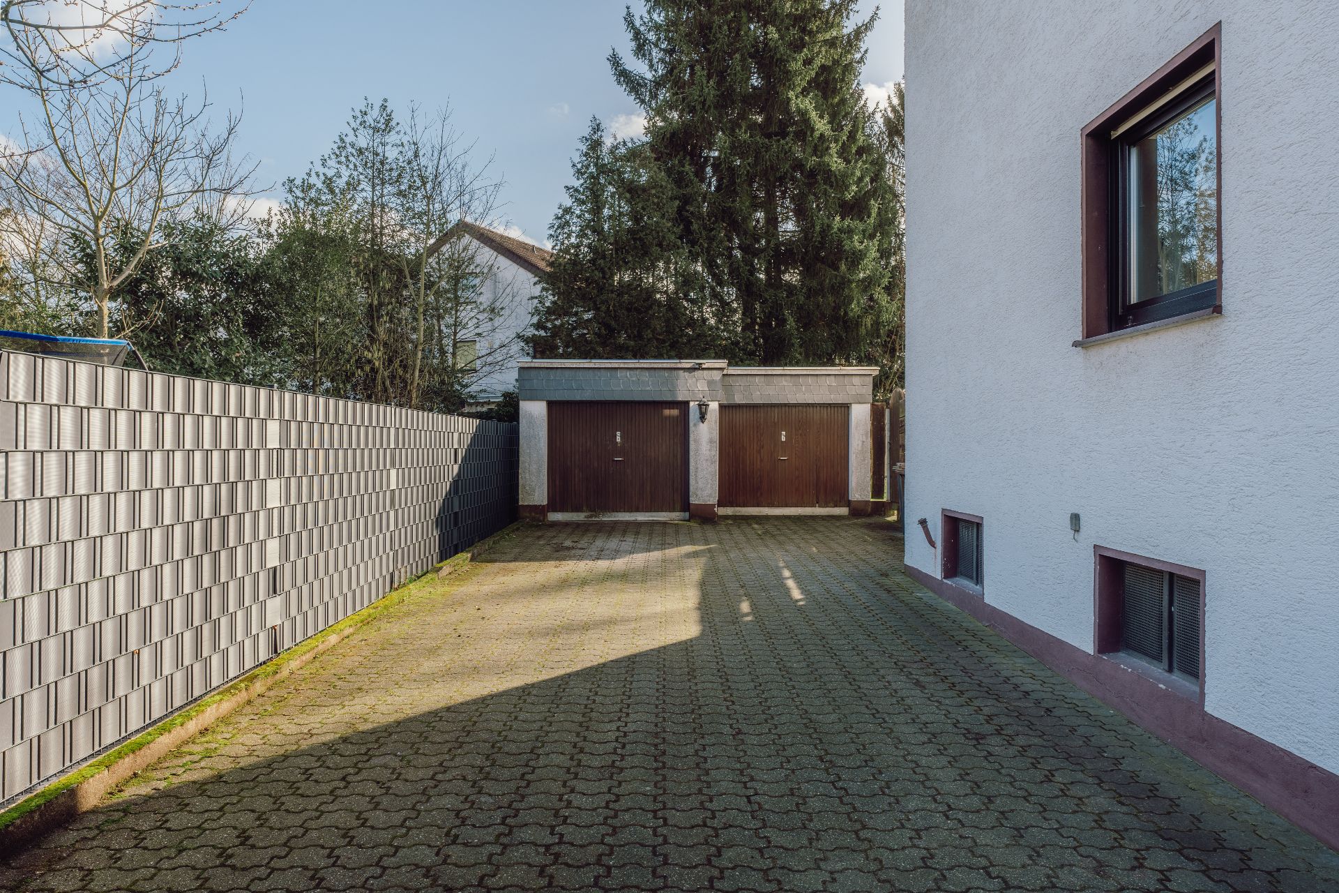 Gepflasterte Einfahrt mit zwei Garagentoren, Metallzaun links und Hausfassade rechts.