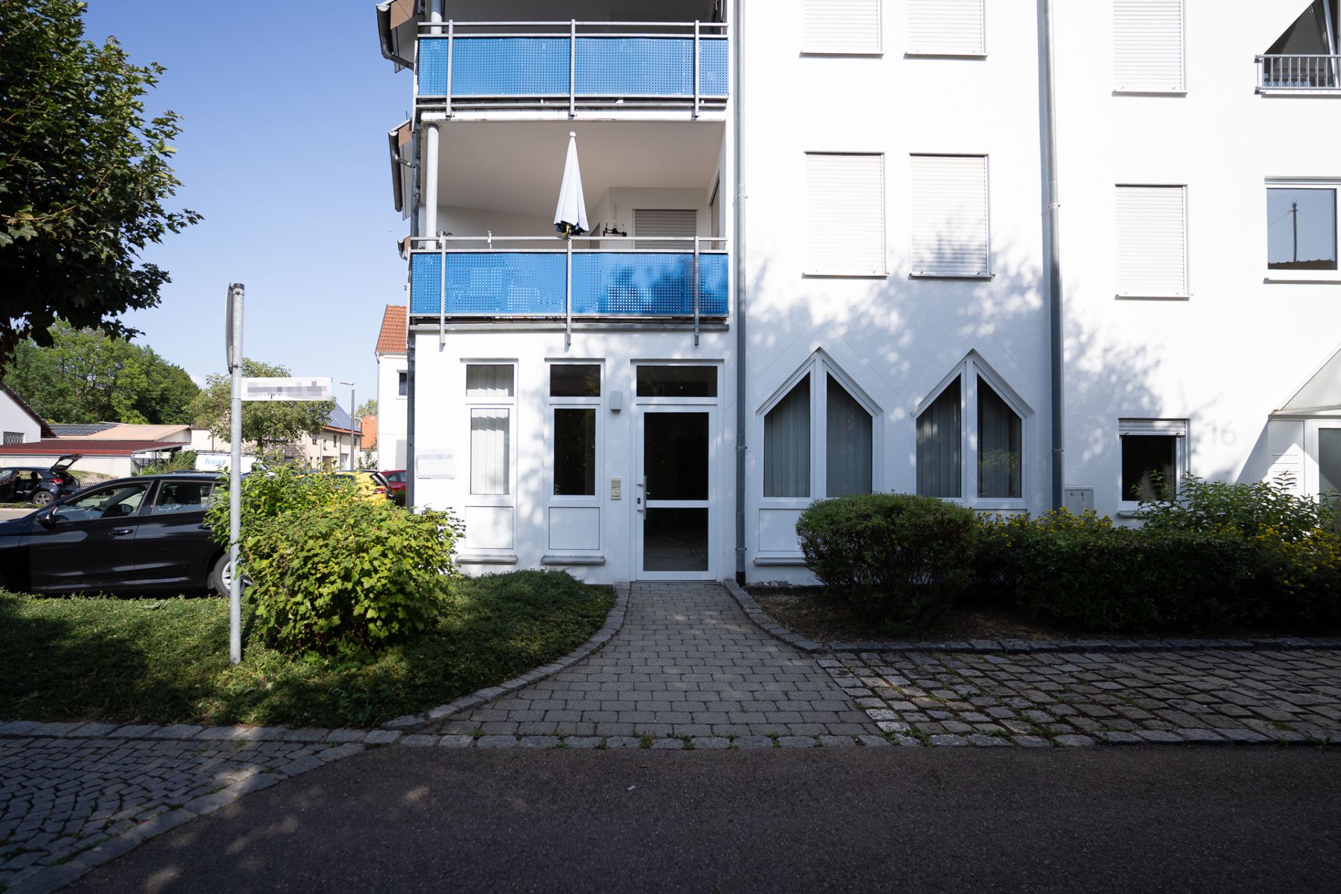 Eingang eines modernen Wohnhauses mit weißen Wänden und blauen Balkonen, umgeben von Büschen.