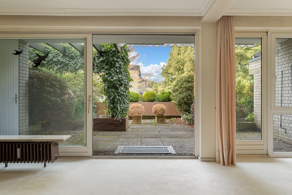 Blick aus einem Wohnzimmer durch große Glasfront auf eine Terrasse mit Pflanzen und Garten.