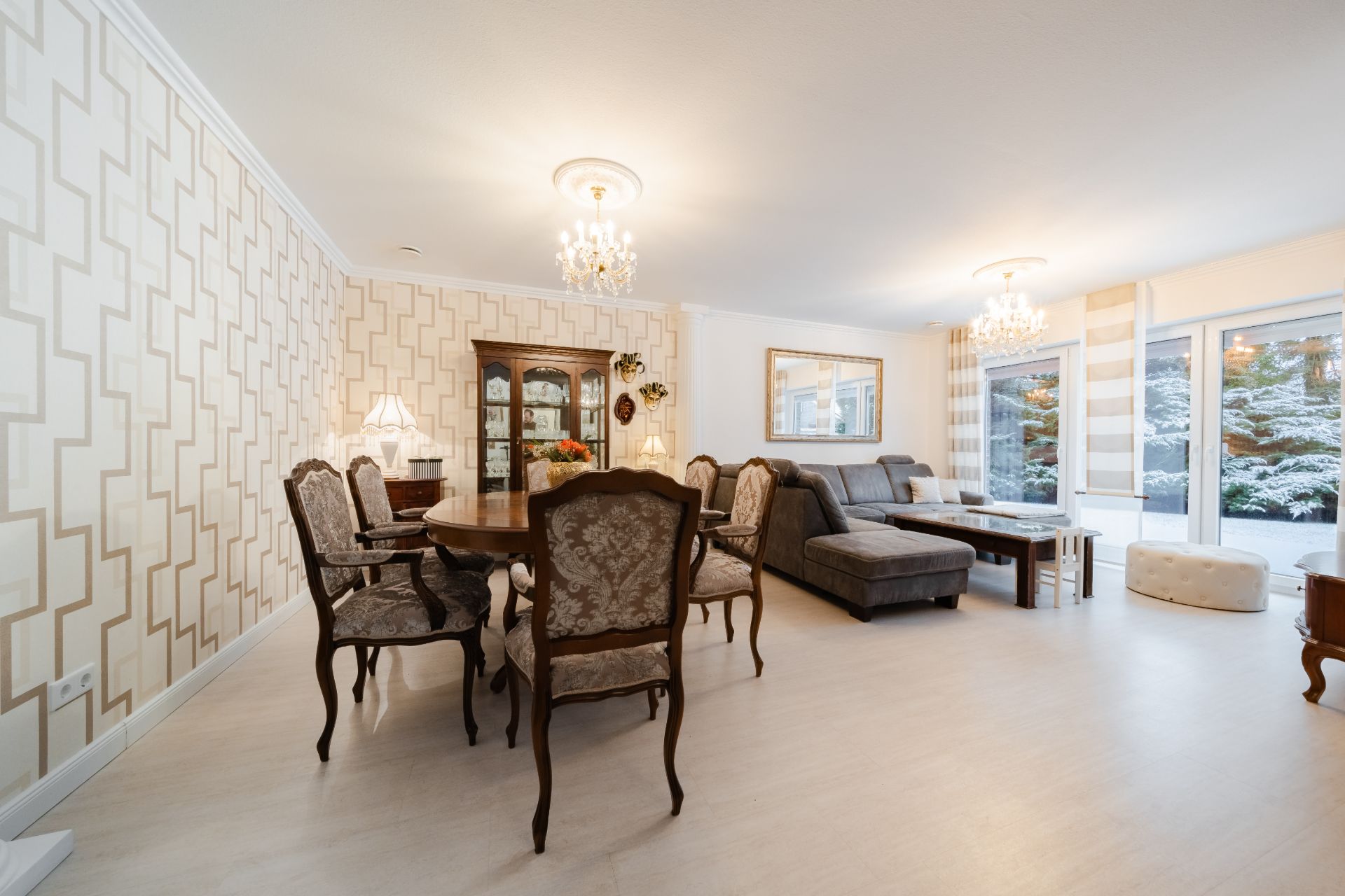 Helles Wohnzimmer mit Esstisch, Stühlen, Sofa, Vitrine und großen Fenstern mit Blick nach draußen.