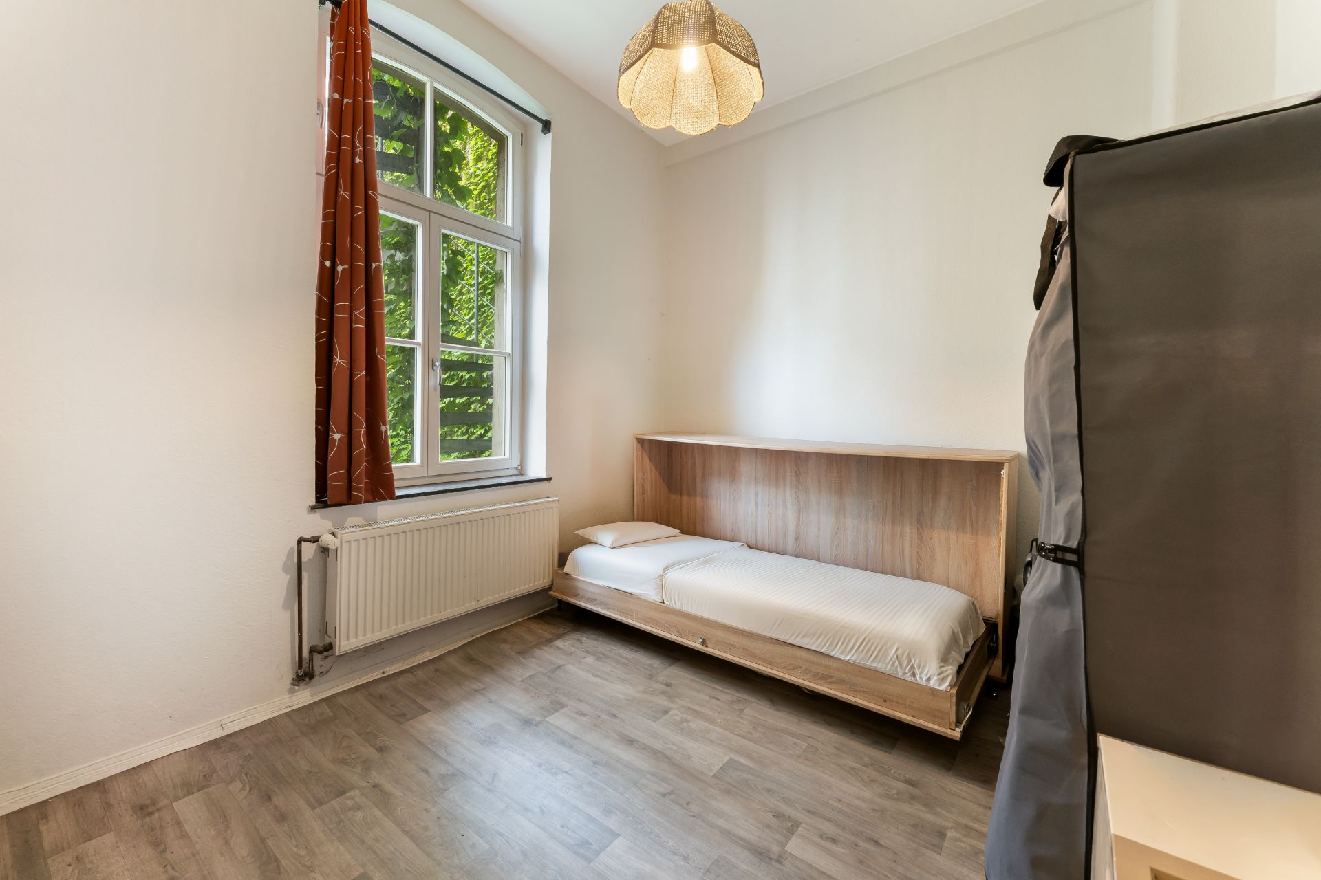 Helles Zimmer mit Einzelbett, großem Fenster, Vorhang und Laminatboden.