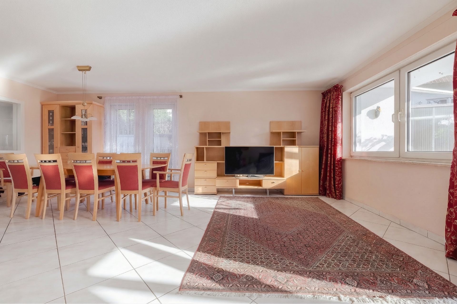 Helles Wohnzimmer mit Esstisch, Stühlen, Fernseher, Regalwand, großen Fenstern und Teppich.