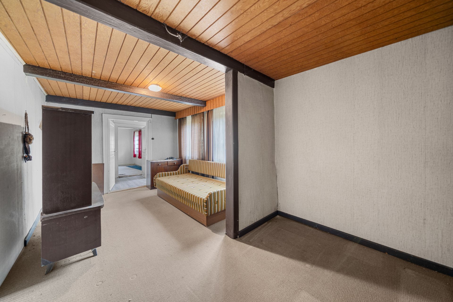 Zimmer mit Holzbalkendecke, Teppichboden, Bett, Kommode und Fenster mit Vorhängen.