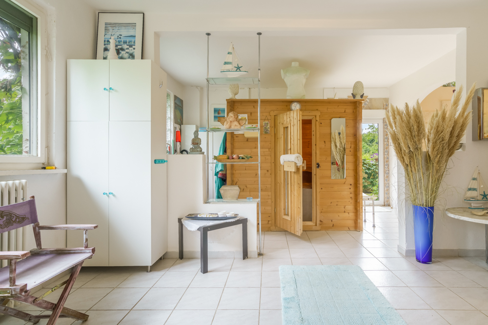 Heller Raum mit Holzsauna, weißem Schrank, Sessel und Deko wie Segelschiffmodellen und Trockenblumen.