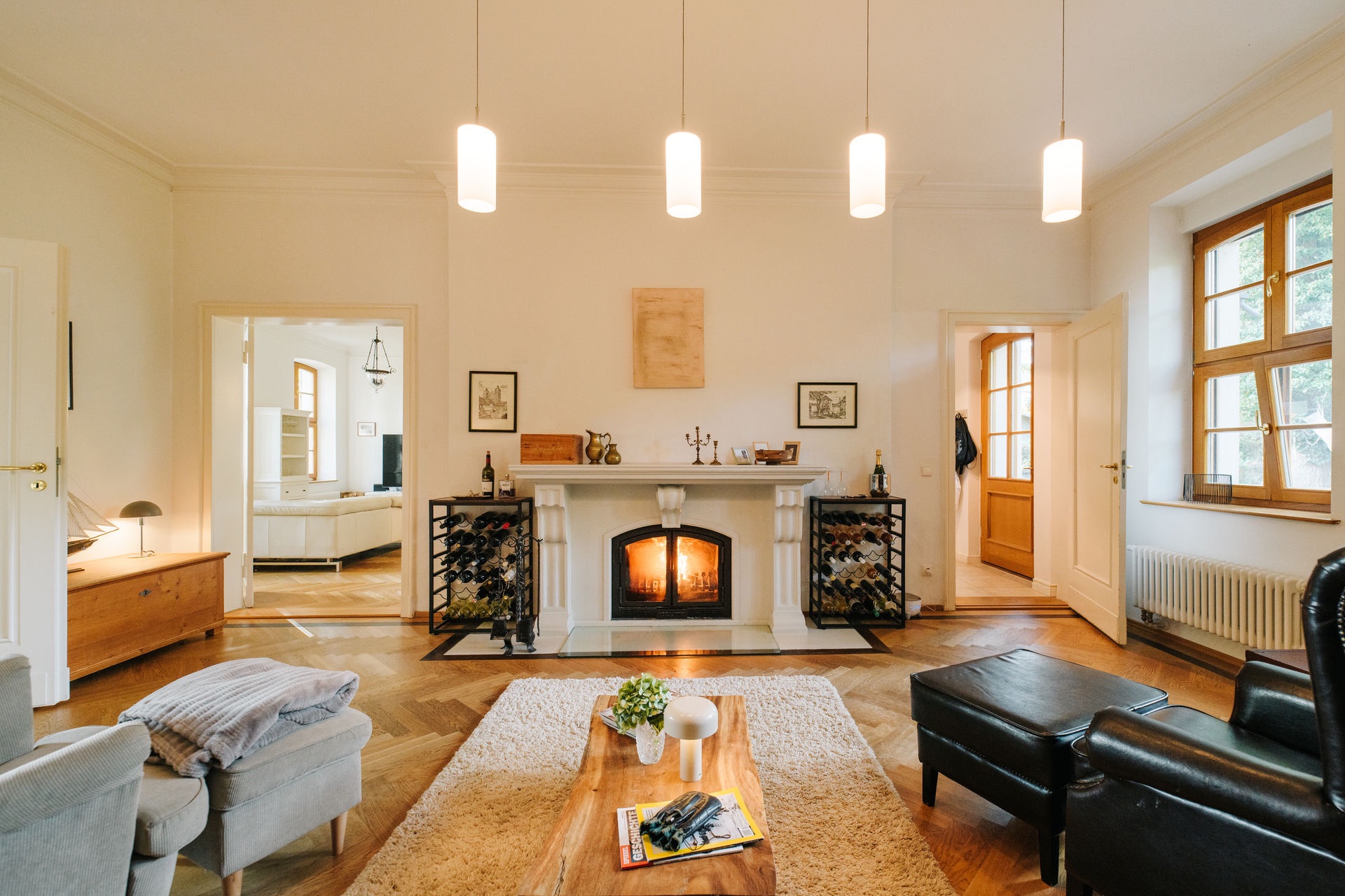 Helles Wohnzimmer mit Kamin, Holzboden, Teppich, Sesseln und großen Fenstern.
