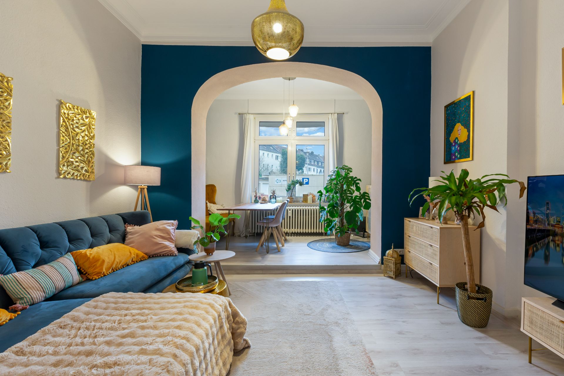 Wohnzimmer mit blauem Sofa, Pflanzen, Holzmöbeln und Blick auf Essbereich mit großem Fenster.