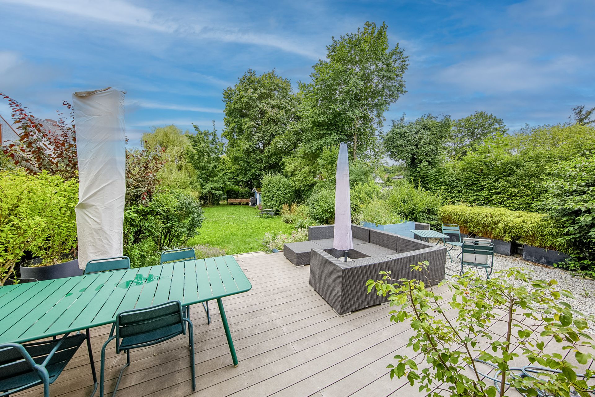 Terrasse mit grünem Tisch, Stühlen und Sitzgruppe, Blick auf Garten mit Bäumen und Büschen.