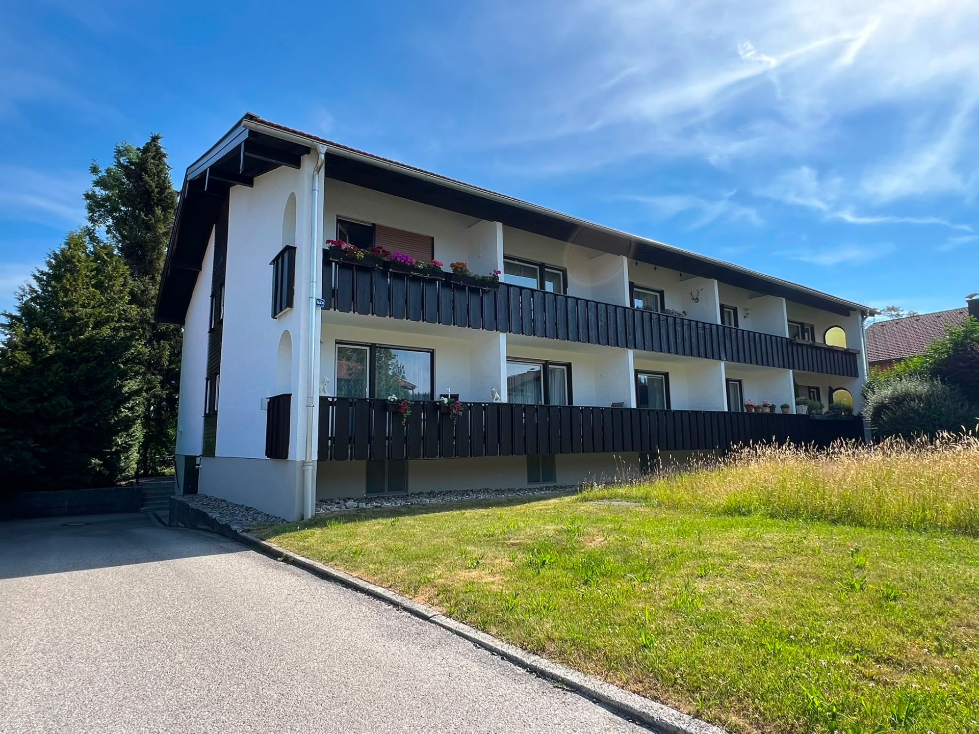 Zweistöckiges Wohnhaus mit Balkonen und Blumen, davor Wiese und Einfahrt.