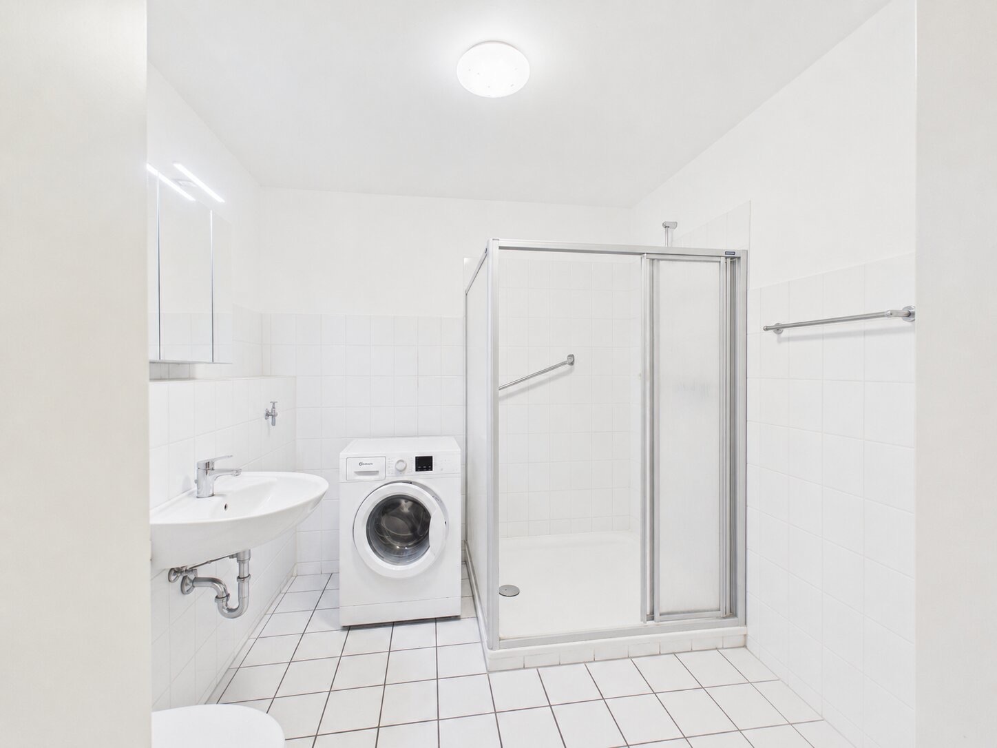 Helles Badezimmer mit Waschbecken, Dusche und Waschmaschine auf gefliestem Boden.