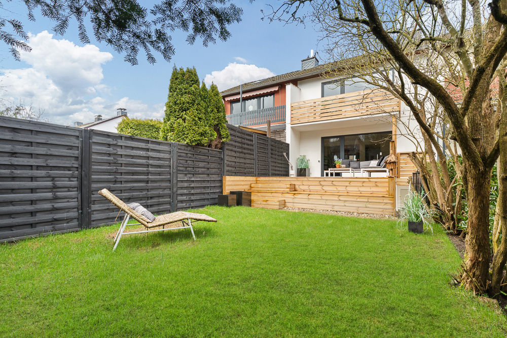 Grüner Garten mit Liege, Holzterrasse und modernem Wohnhaus im Hintergrund.