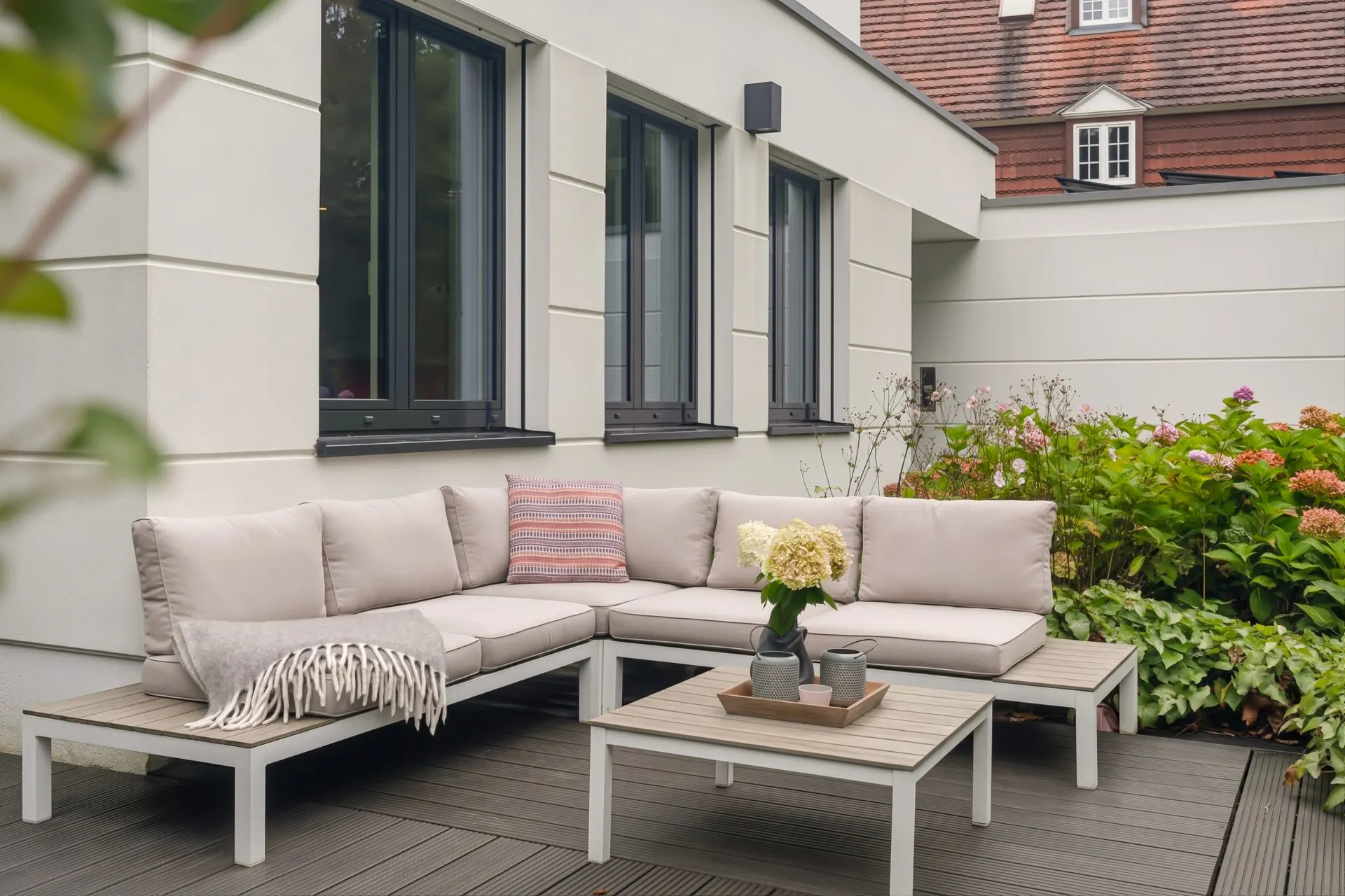 Moderne Terrasse mit Ecksofa, kleinem Tisch, Kissen und Pflanzen neben einem Wohnhaus.