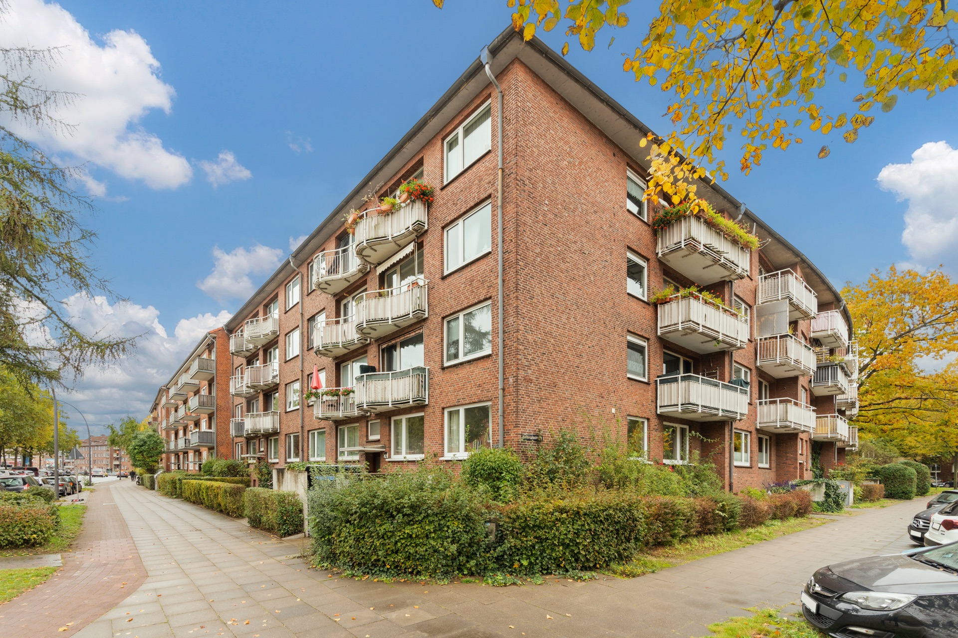 Rotes Mehrfamilienhaus mit Balkonen und grünen Büschen an einer Straßenecke.