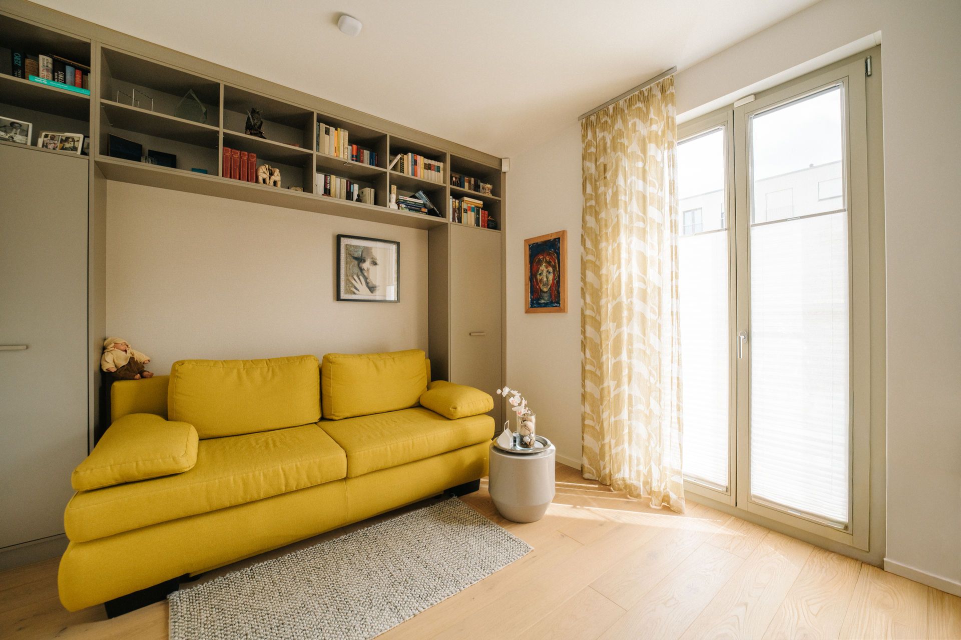 Wohnzimmer mit gelbem Sofa, Regal mit Büchern, rundem Tisch und großen Fenstern mit hellen Vorhängen.