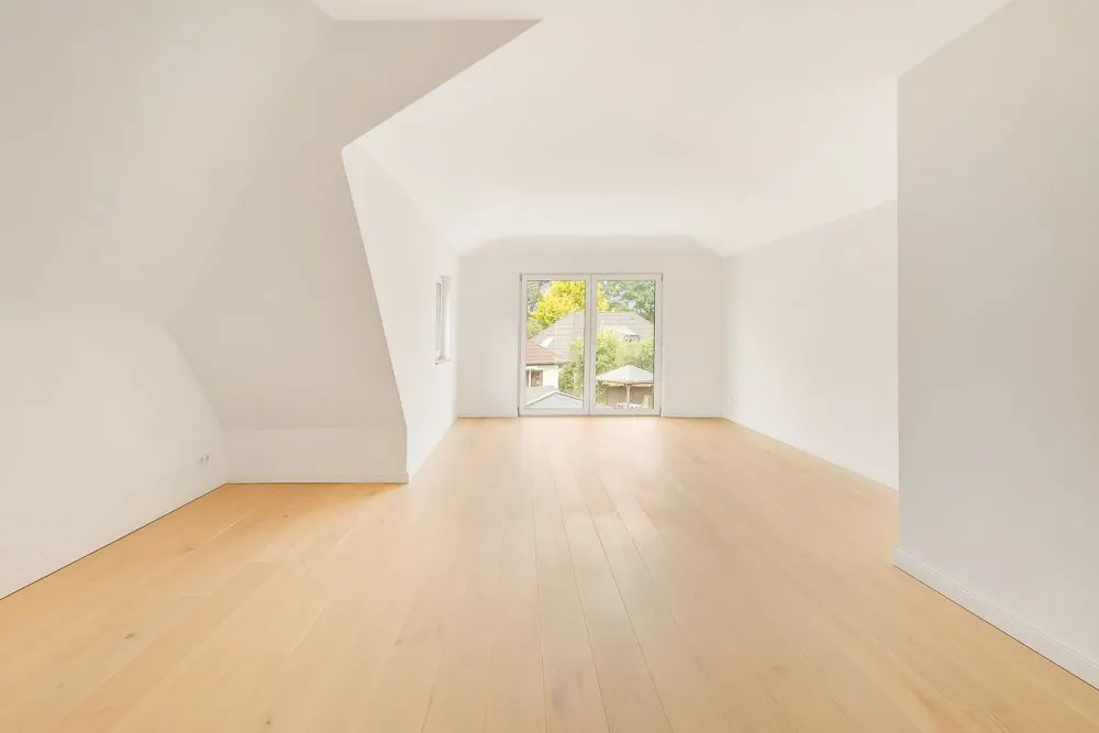 Heller leerer Raum mit Holzfußboden, weißen Wänden und großem Fenster zur Terrasse.