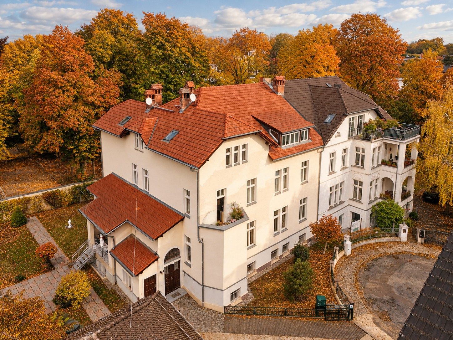 Großes, helles Wohnhaus mit rotem Dach, umgeben von Bäumen im Herbst.