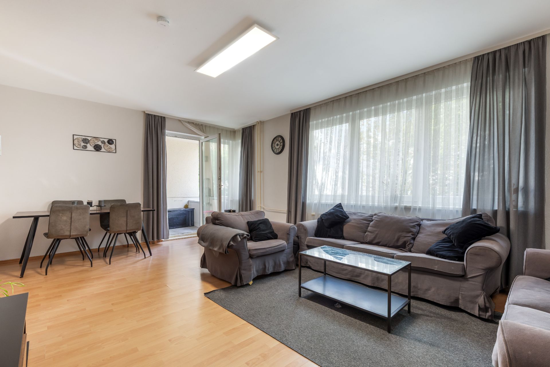 Wohnzimmer mit grauem Sofa, Sessel, Couchtisch und Essbereich, große Fenster mit Vorhängen.