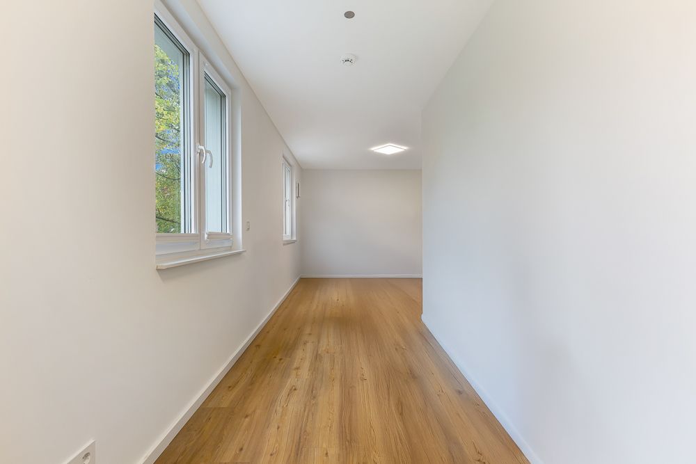 Schmaler, leerer Raum mit hellem Holzboden und zwei Fenstern an der linken Wand.