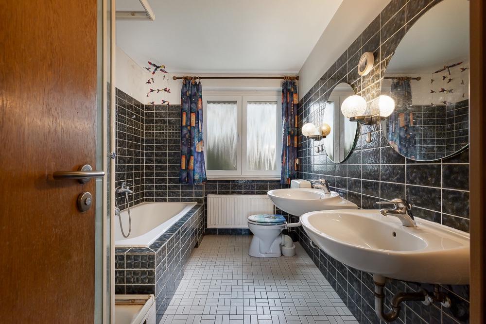 Badezimmer mit zwei Waschbecken, Badewanne, Toilette und zwei Fenstern mit blauen Vorhängen.