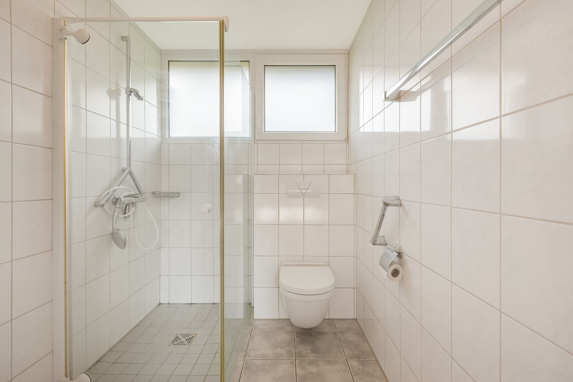Modernes Bad mit Dusche, WC und zwei Fenstern, alles mit weißen Fliesen ausgestattet.
