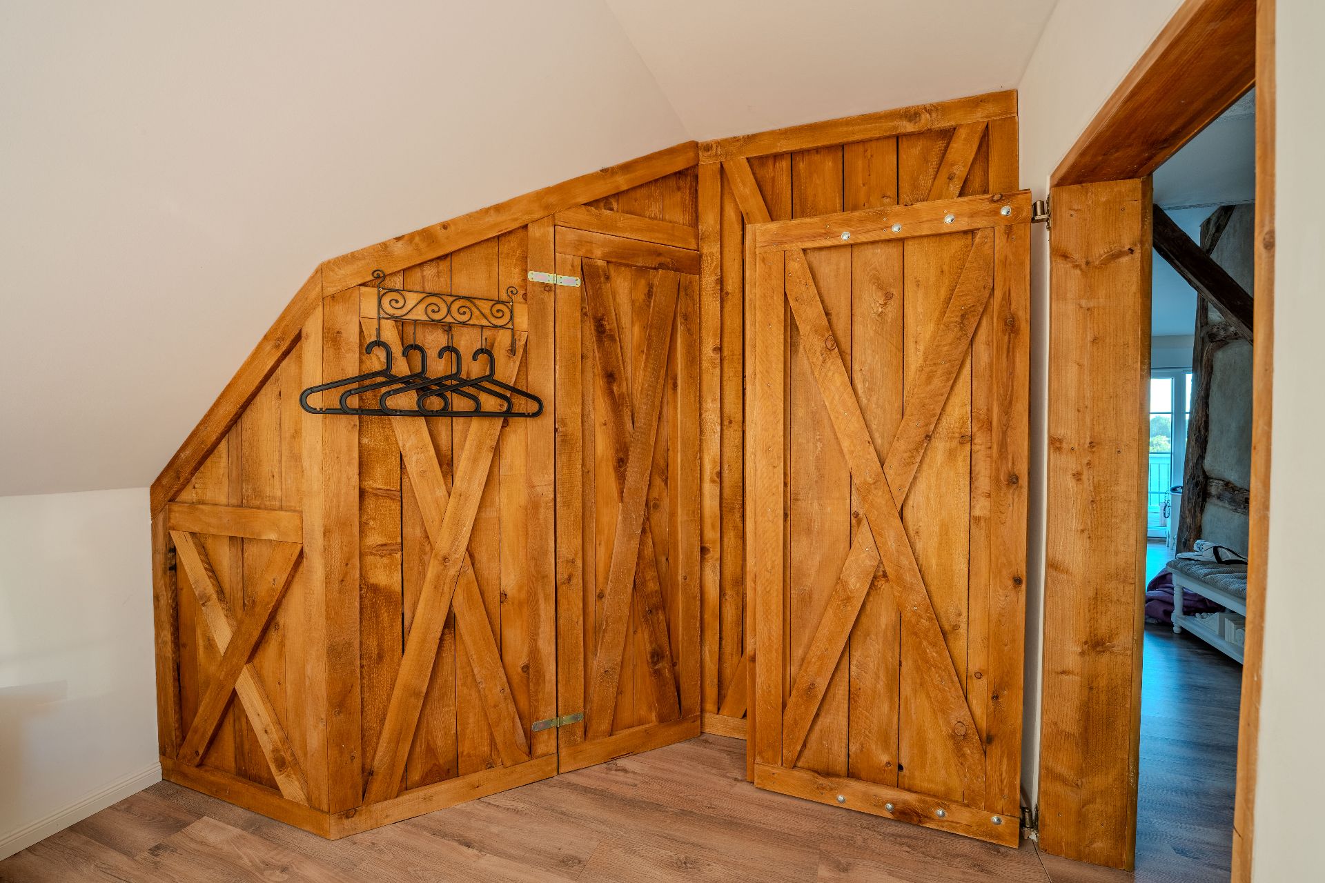 Holzschrank mit offenen Türen und Kleiderbügeln an einer Dachschräge.