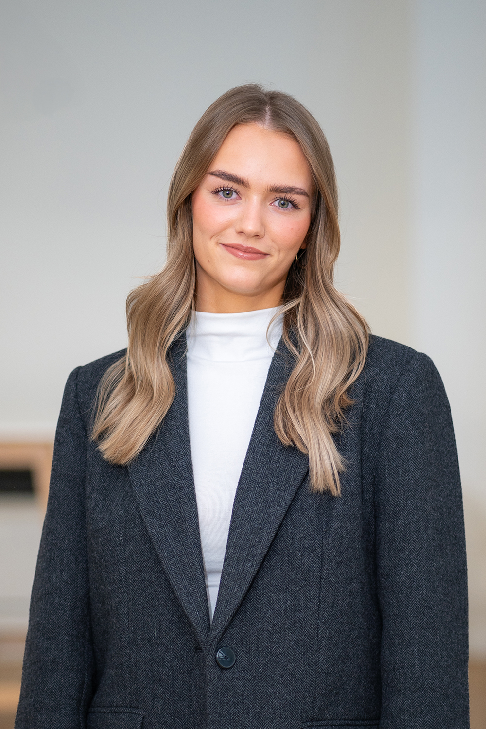 Junge Frau mit langen blonden Haaren in dunklem Blazer vor hellem Hintergrund.