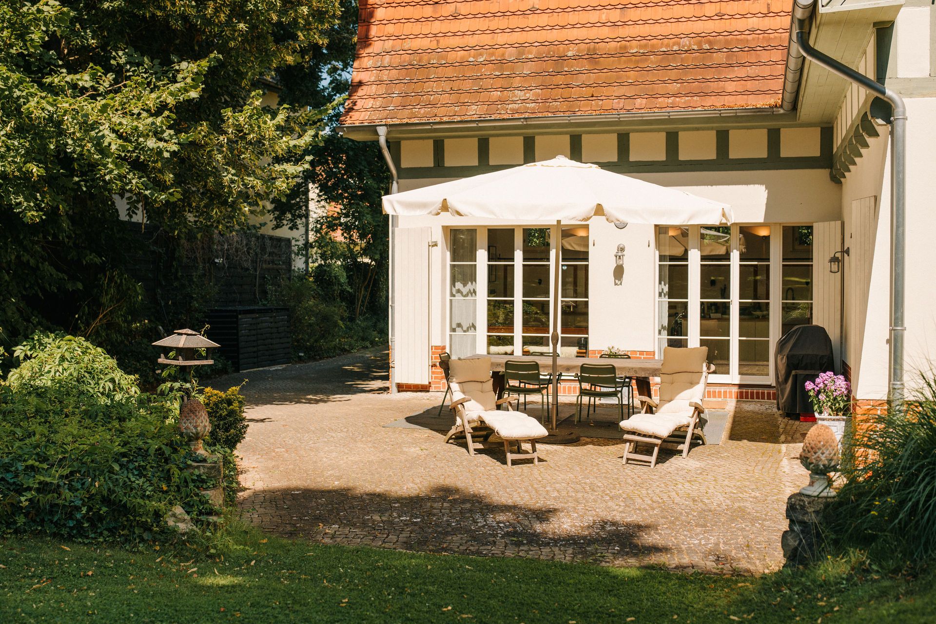 Sonnige Terrasse mit Sitzgruppe, Sonnenliegen, Sonnenschirm und Gartenblick an einem Wohnhaus.