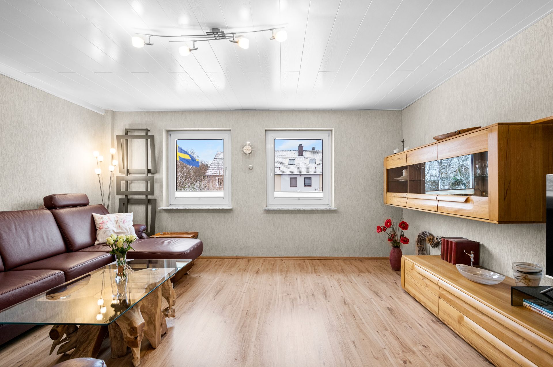 Helles Wohnzimmer mit braunem Sofa, Glastisch, Holzschränken und zwei Fenstern.