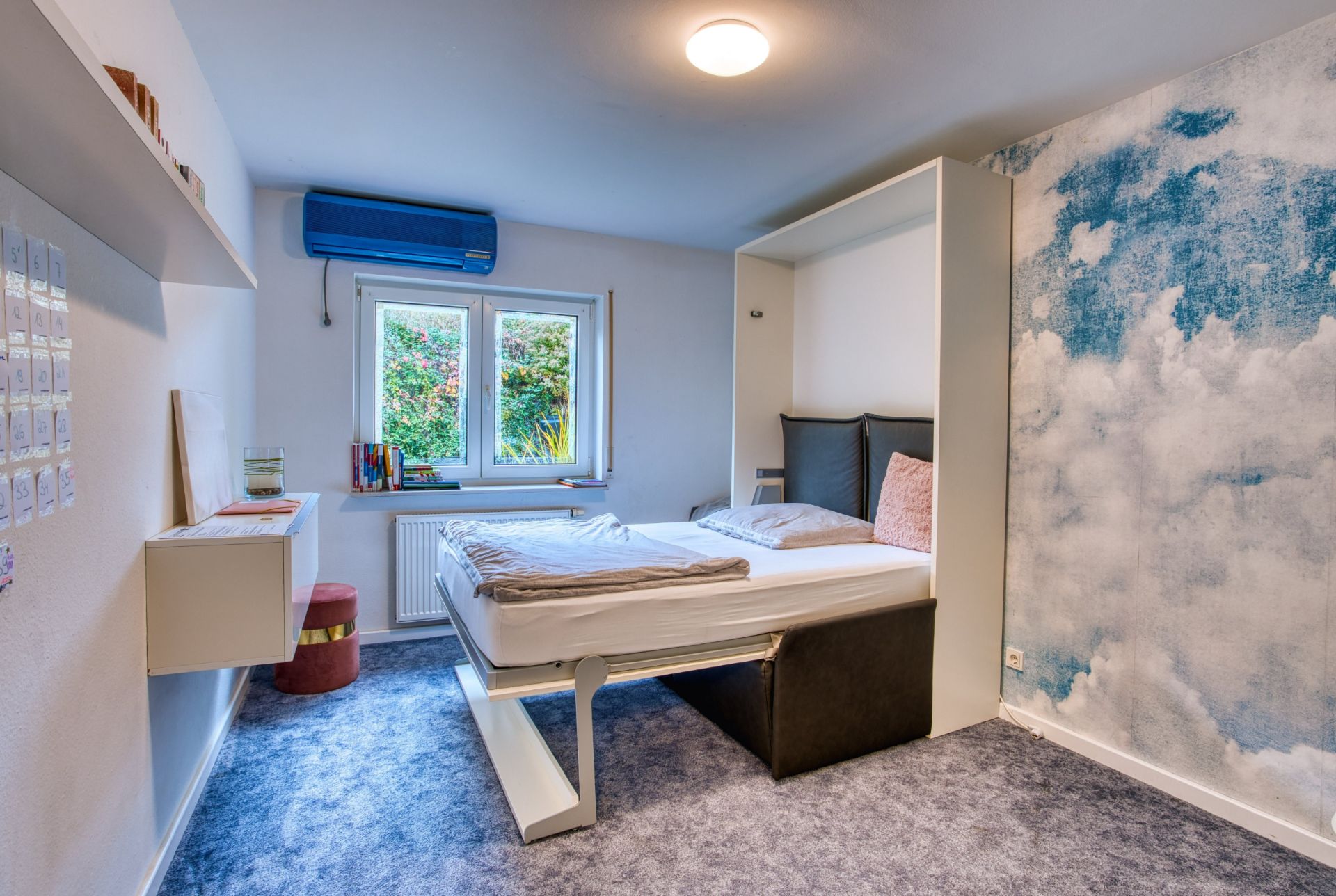 Helles Schlafzimmer mit Ausklappbett, Teppichboden und einer Wand mit Wolkenmotiv.