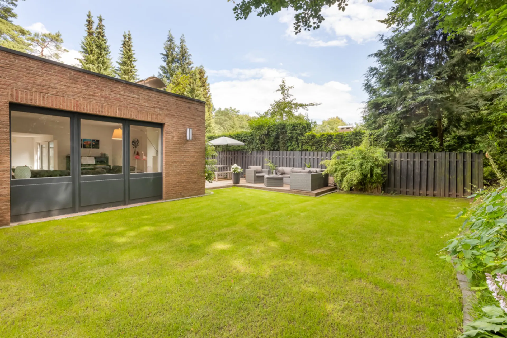 Moderner Garten mit Terrasse, Sitzgruppe und großem Rasen an einem Haus mit großen Fenstern.