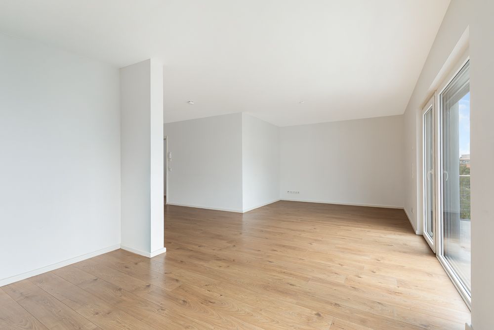 Heller, leerer Raum mit Holzfußboden und großen Fenstern auf der rechten Seite.