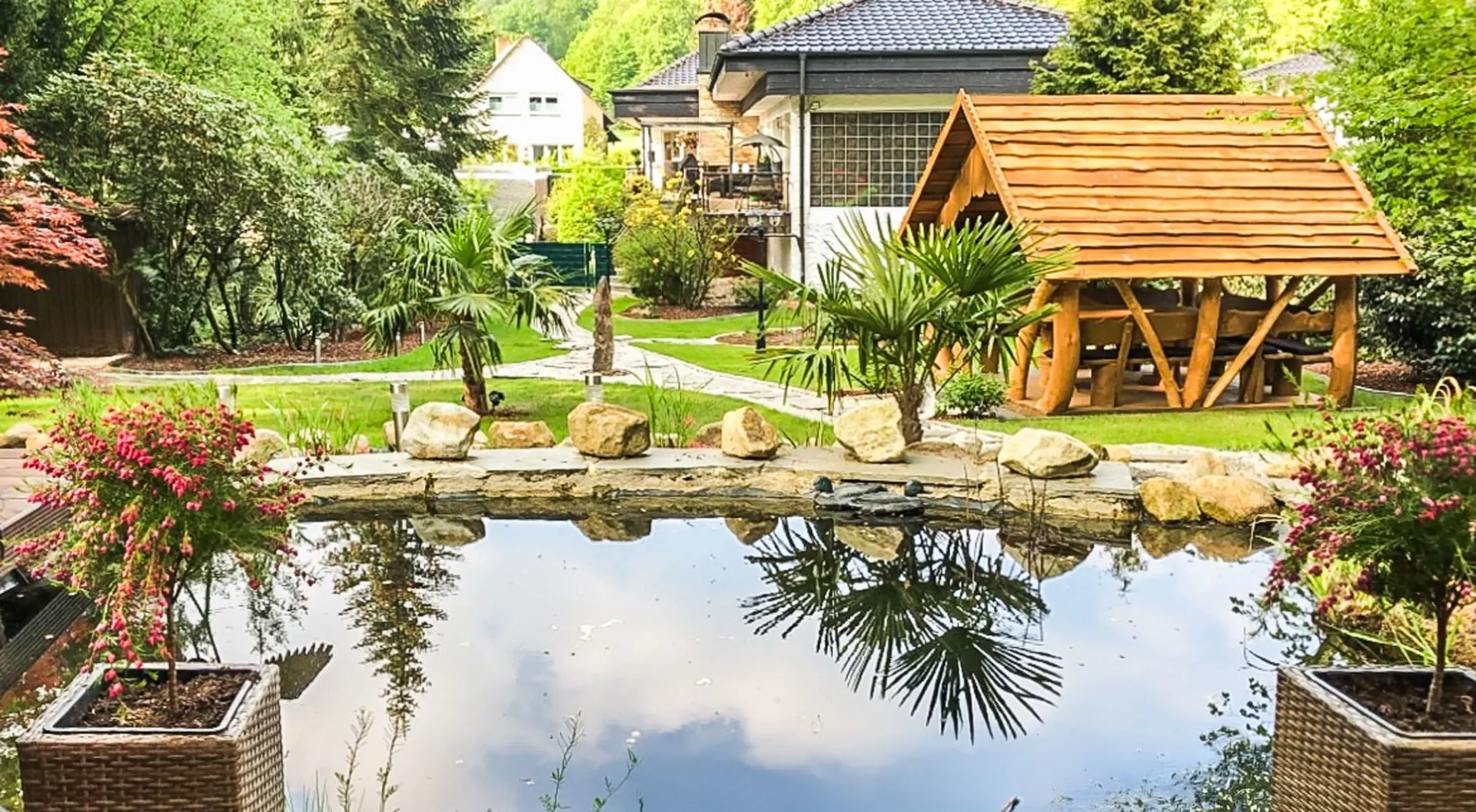 Garten mit kleinem Teich, Steinen, Pflanzen und Holzpavillon vor einem Wohnhaus.