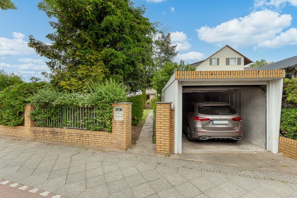 Einfahrt mit Garage, Auto steht in der Garage, daneben Gartenzaun mit Briefkasten und viel Grün.