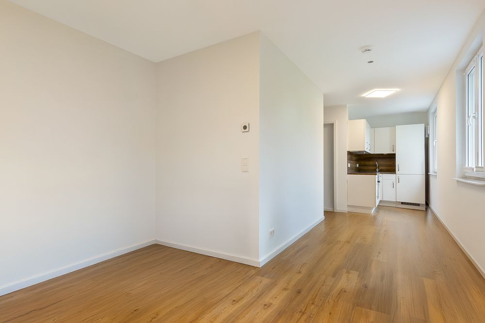 Leerer Raum mit Holzboden und Zugang zu einer weißen Küche mit Fenstern.