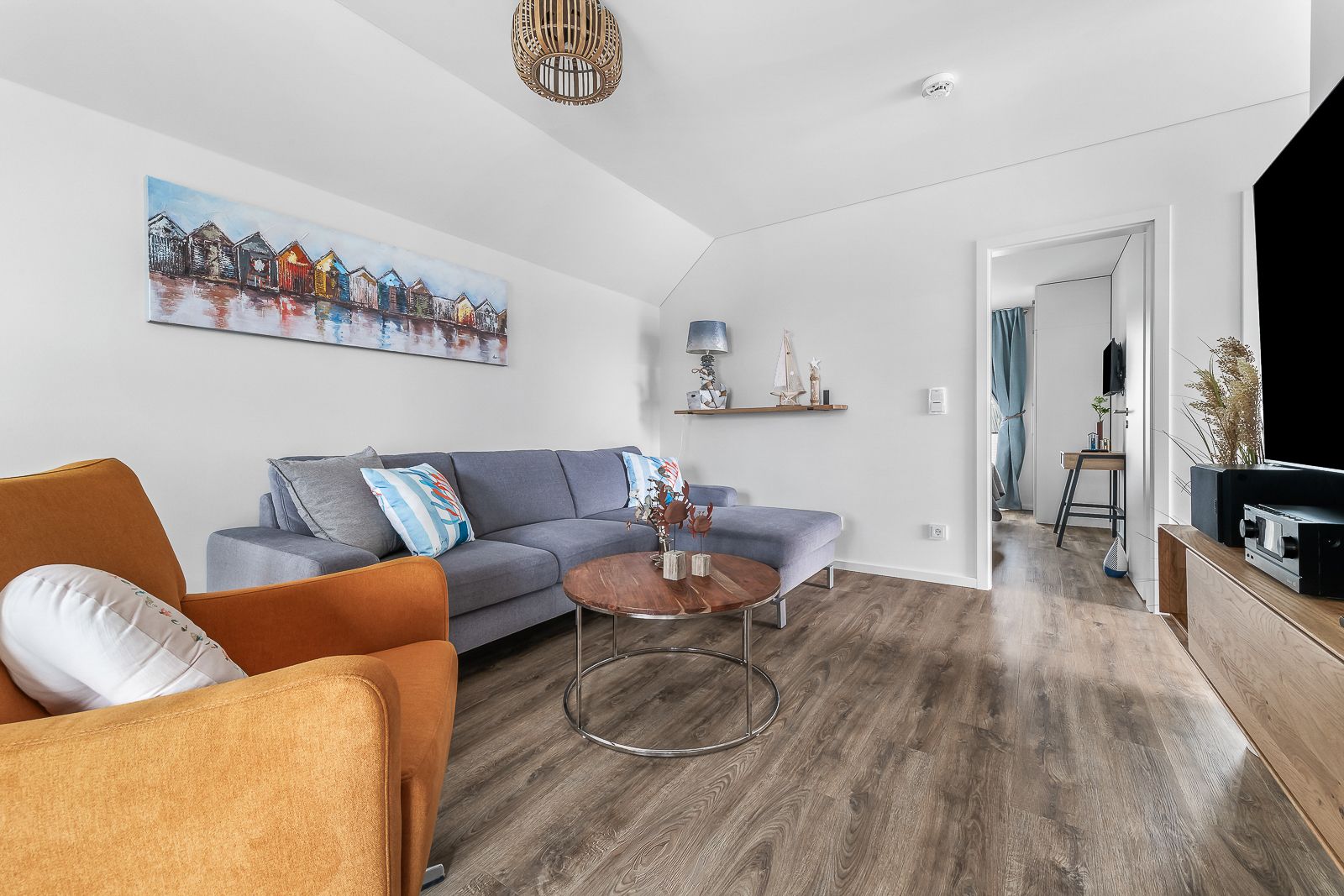 Wohnzimmer mit grauem Ecksofa, orangefarbenem Sessel und rundem Holztisch auf Holzfußboden.