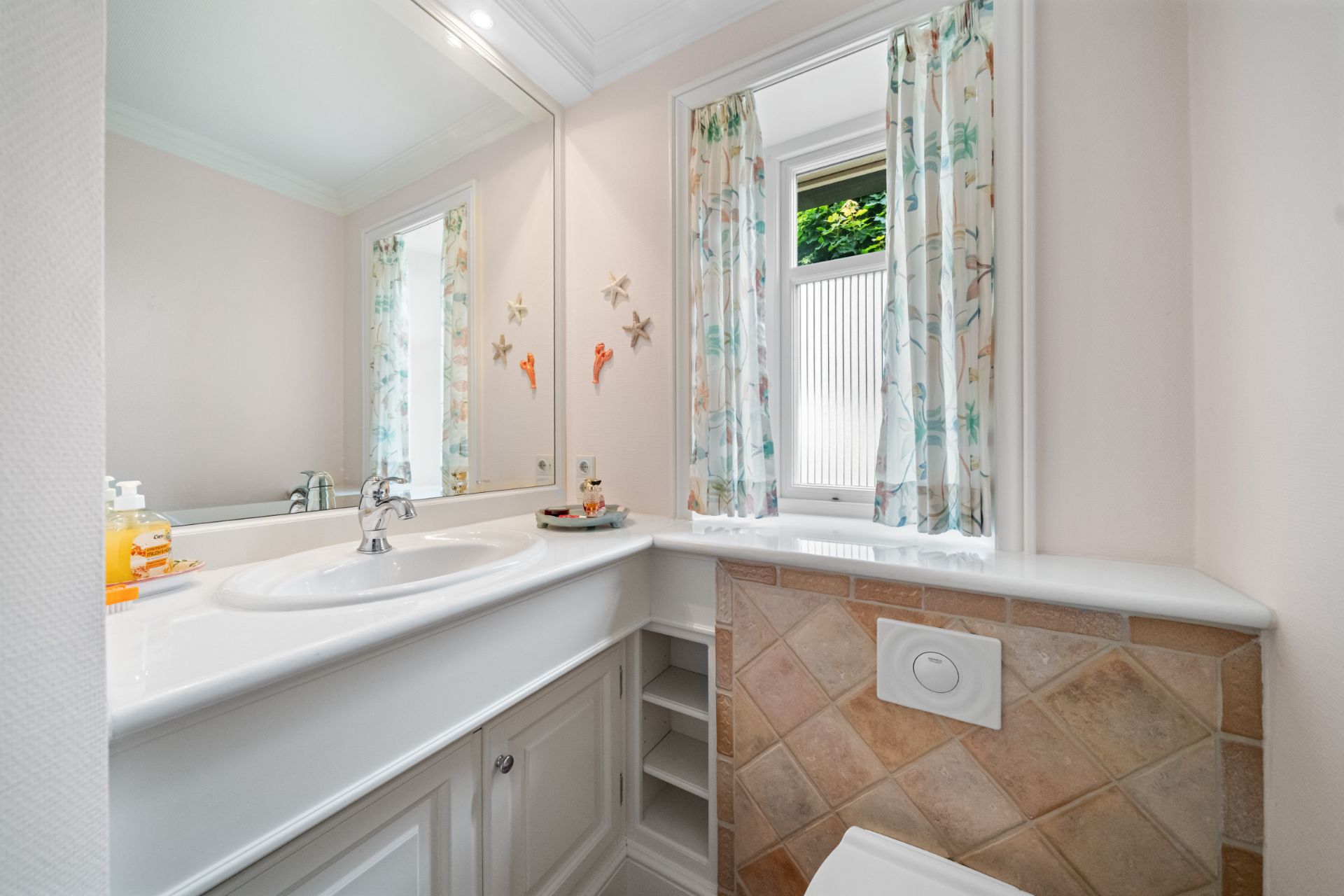Helles Badezimmer mit großem Spiegel, Waschbecken, WC und Fenster mit bunten Vorhängen.
