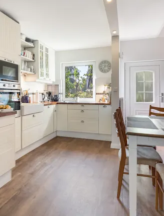 Helle Küche mit Holzfußboden, Esstisch, Stühlen und Treppe. Obstkorb und Blumen sind sichtbar.