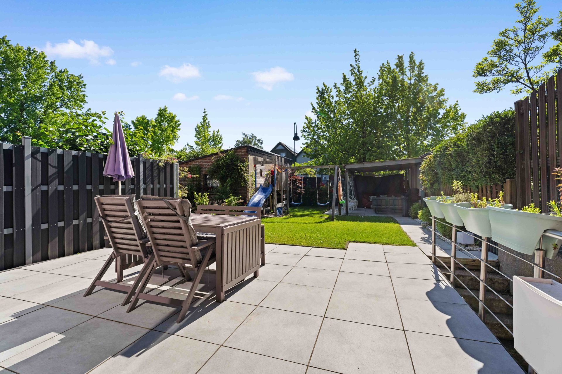 Gepflasterte Terrasse mit Holztisch, Stühlen und Garten, umgeben von Bäumen und einem Holzzaun.