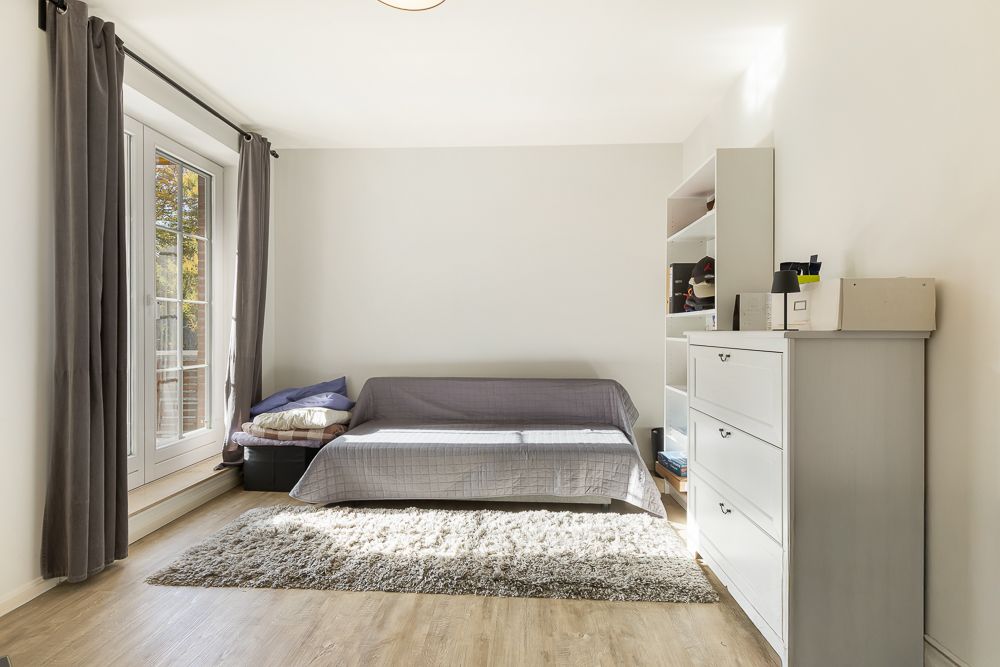 Helles Zimmer mit grauem Sofa, Teppich, Kommode und Regal, Fenster mit grauen Vorhängen.