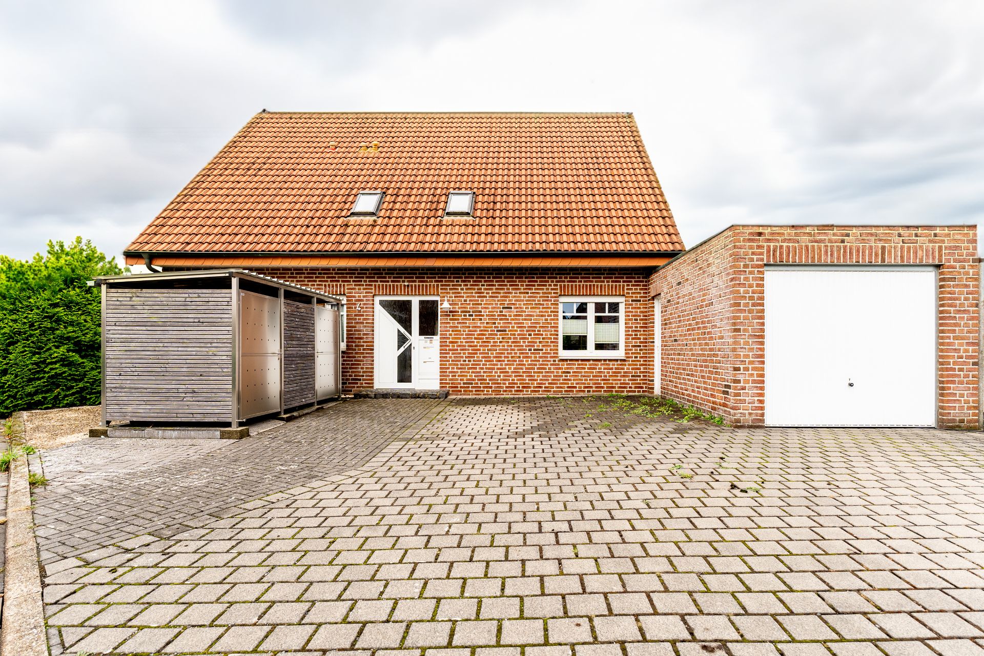 Backsteinhaus mit rotem Dach, weißer Tür, Garage und gepflastertem Hof.