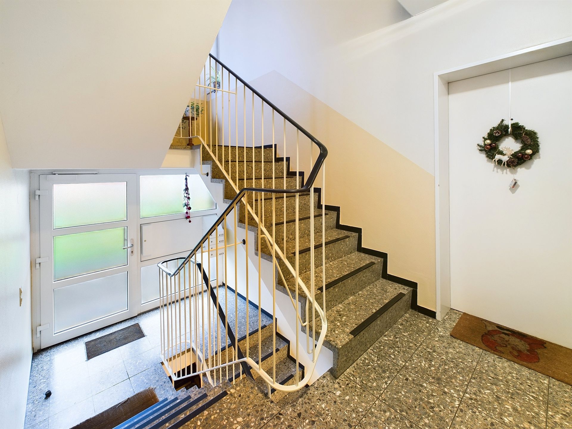 Helles Treppenhaus mit Geländer, Glastür und dekoriertem Wohnungseingang.