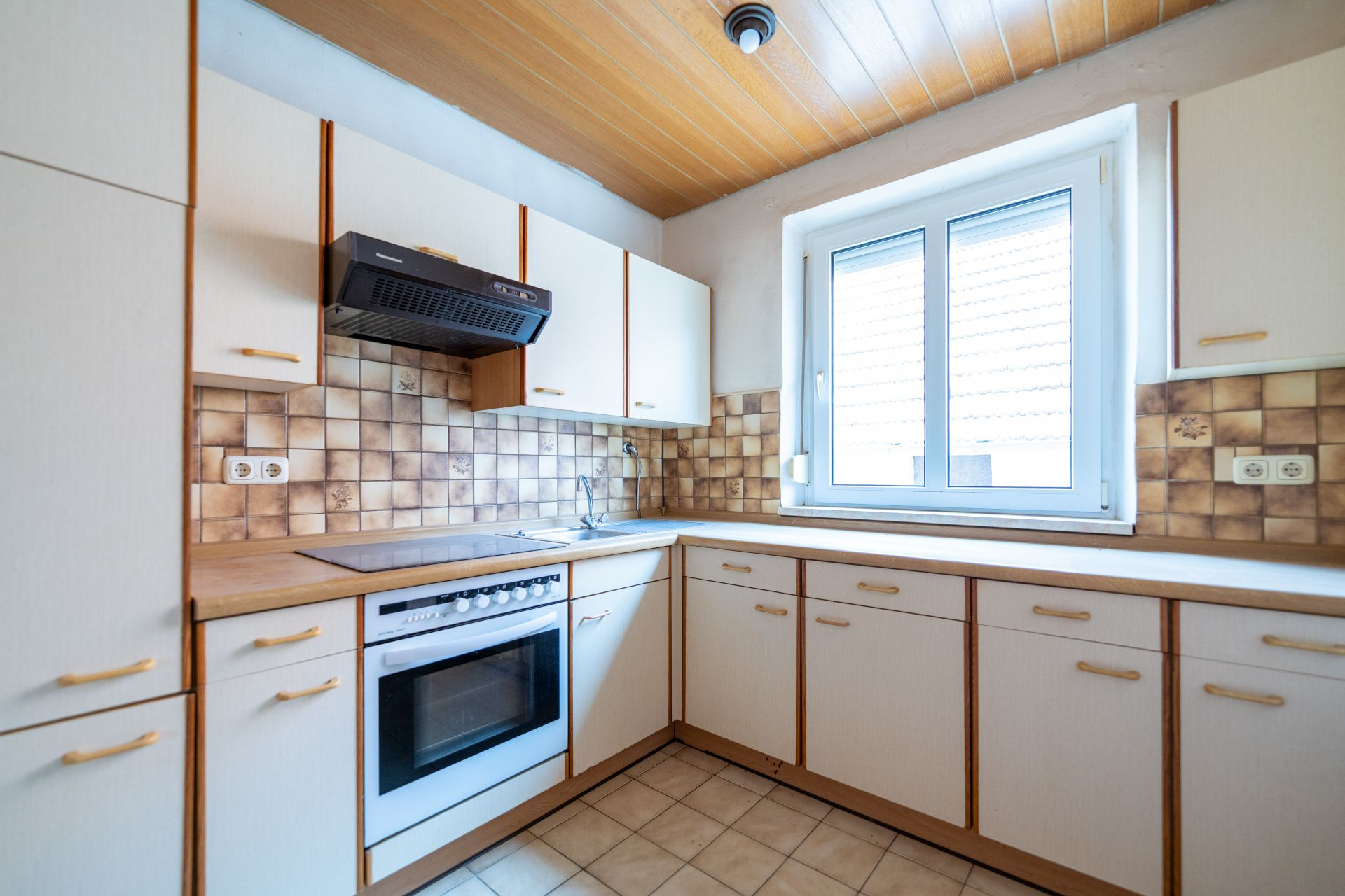 Helle Einbauküche mit Herd, Spüle, weißen Schränken und Fenster, braune Fliesen an der Wand.