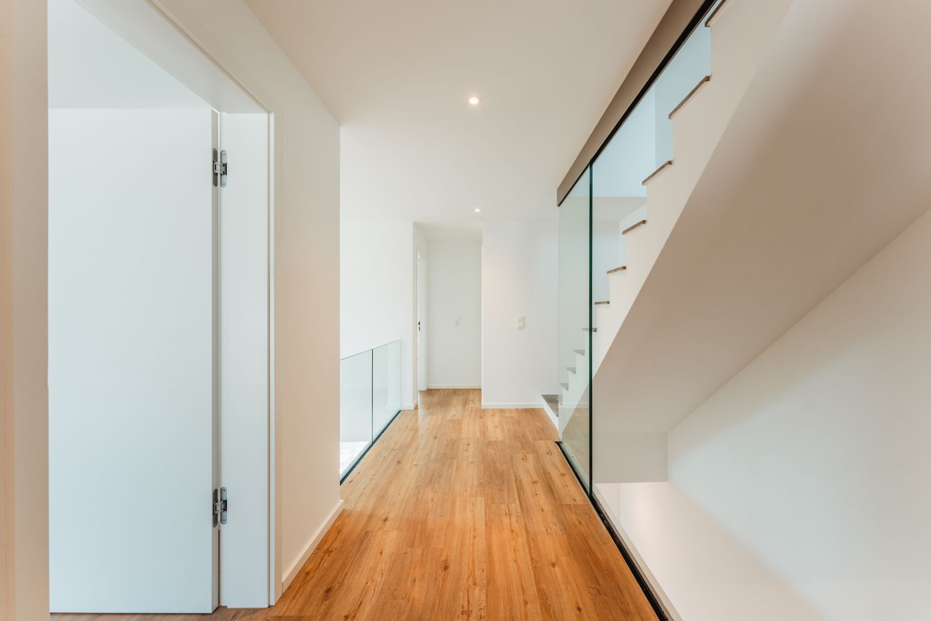 Heller Flur mit Holzboden, weißen Wänden und Treppe mit Glasgeländer.