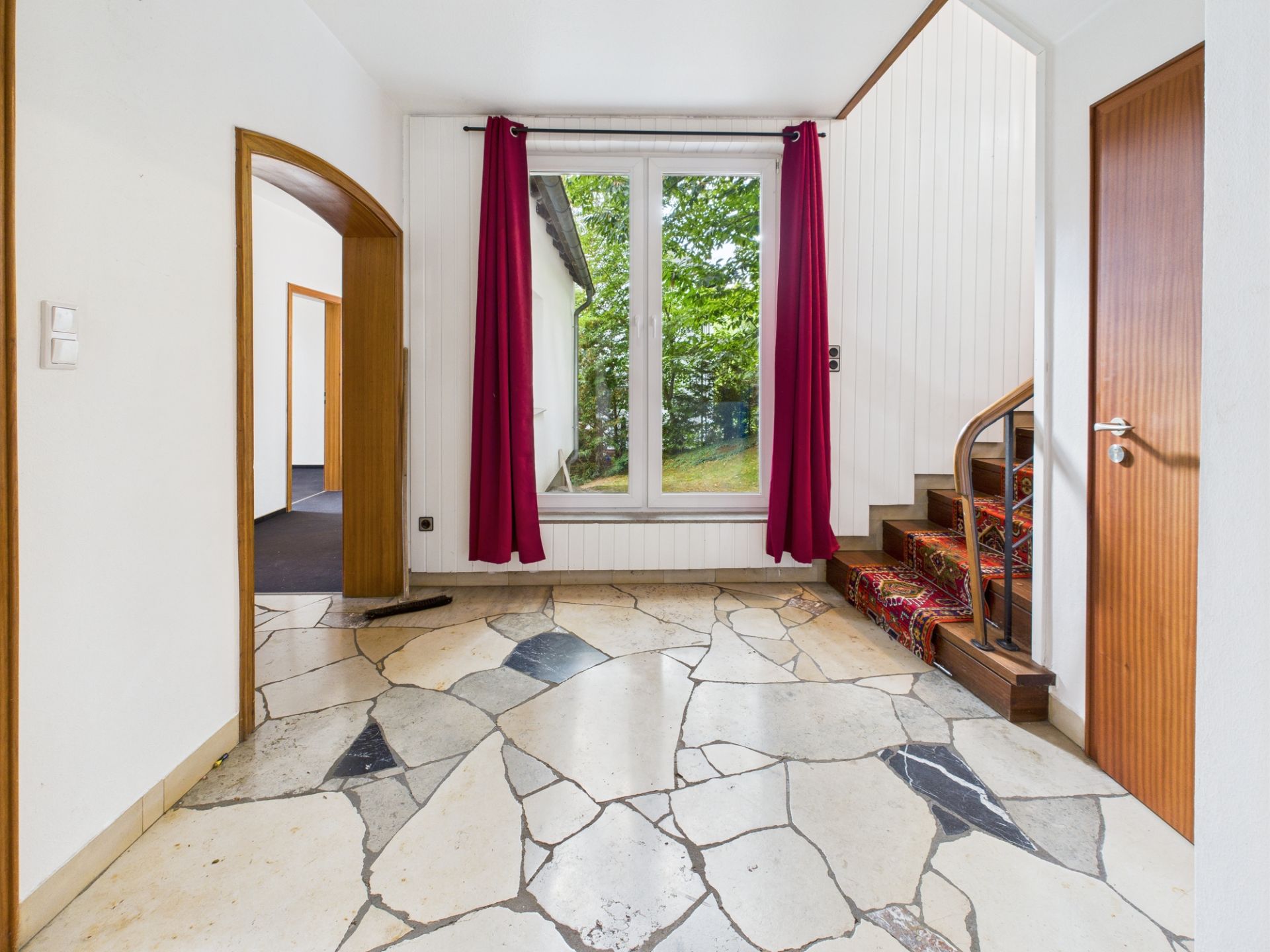 Flur mit Natursteinboden, Treppe, großem Fenster und roten Vorhängen.