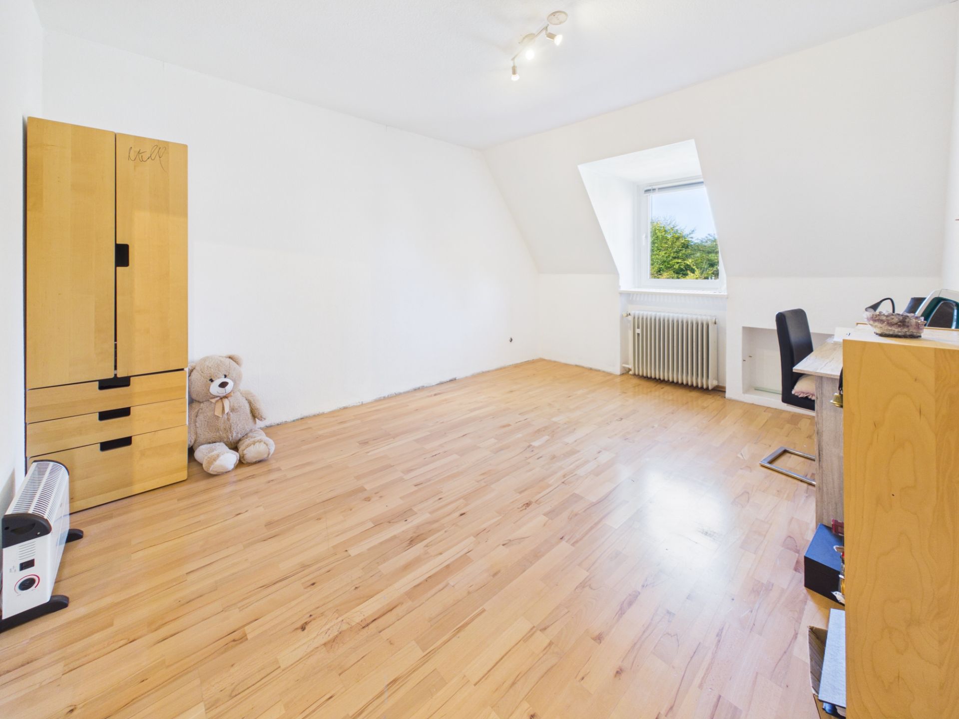 Helles Zimmer mit Holzfußboden, Schrank, großem Teddybär, Schreibtisch und Fenster.