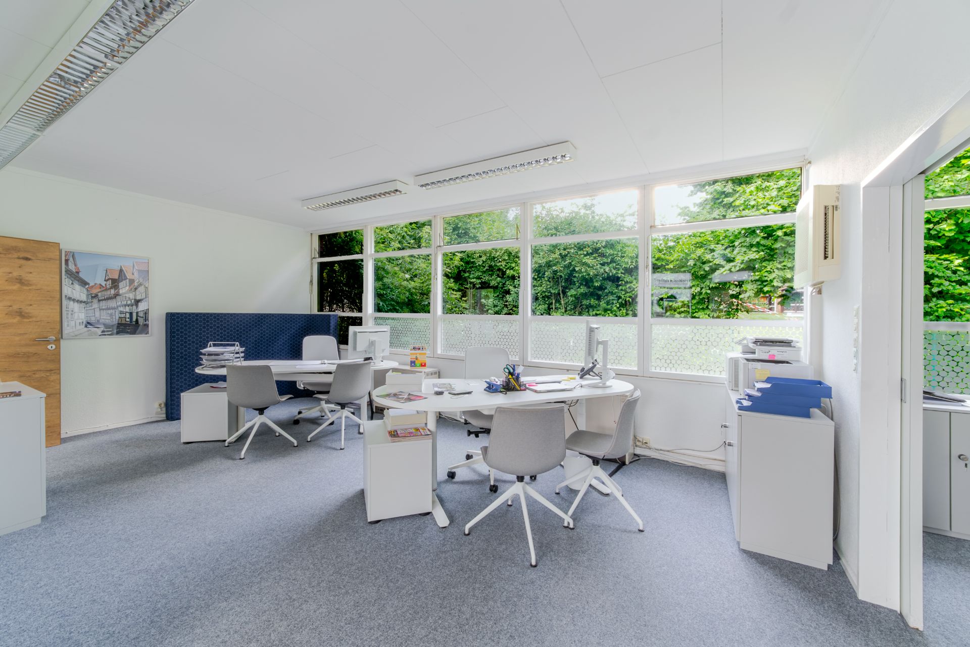 Helles Büro mit mehreren Schreibtischen, Bürostühlen und großen Fenstern zum Garten.