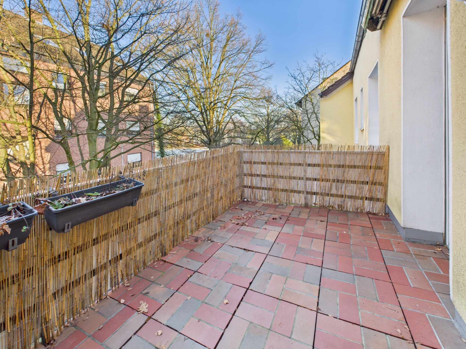Balkon mit roten Bodenfliesen, Bambus-Sichtschutz und Blumenkästen an der Brüstung.