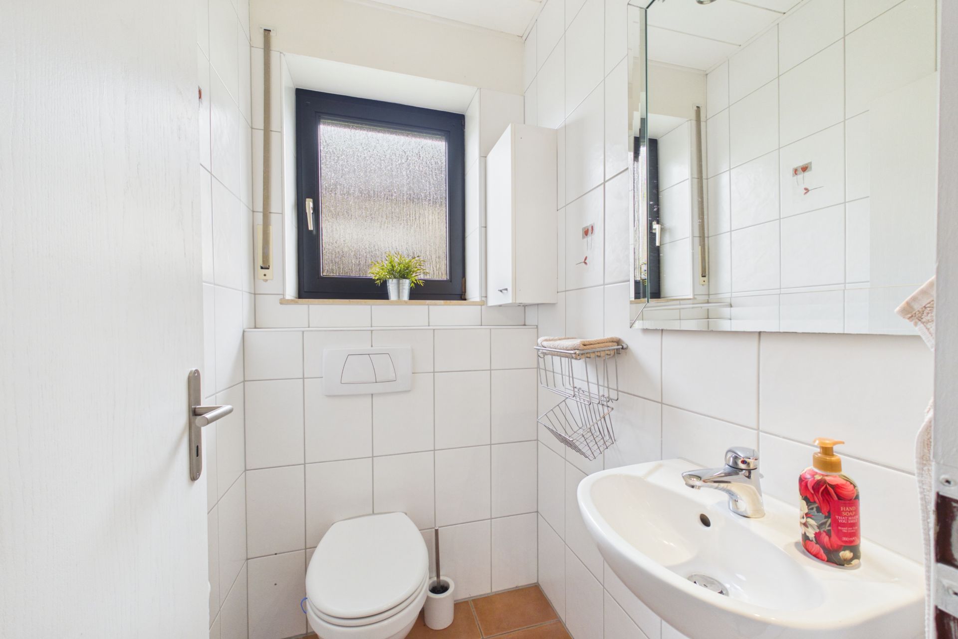 Kleines Badezimmer mit WC, Waschbecken, Spiegel und Fenster. Seifenspender steht am Waschbecken.